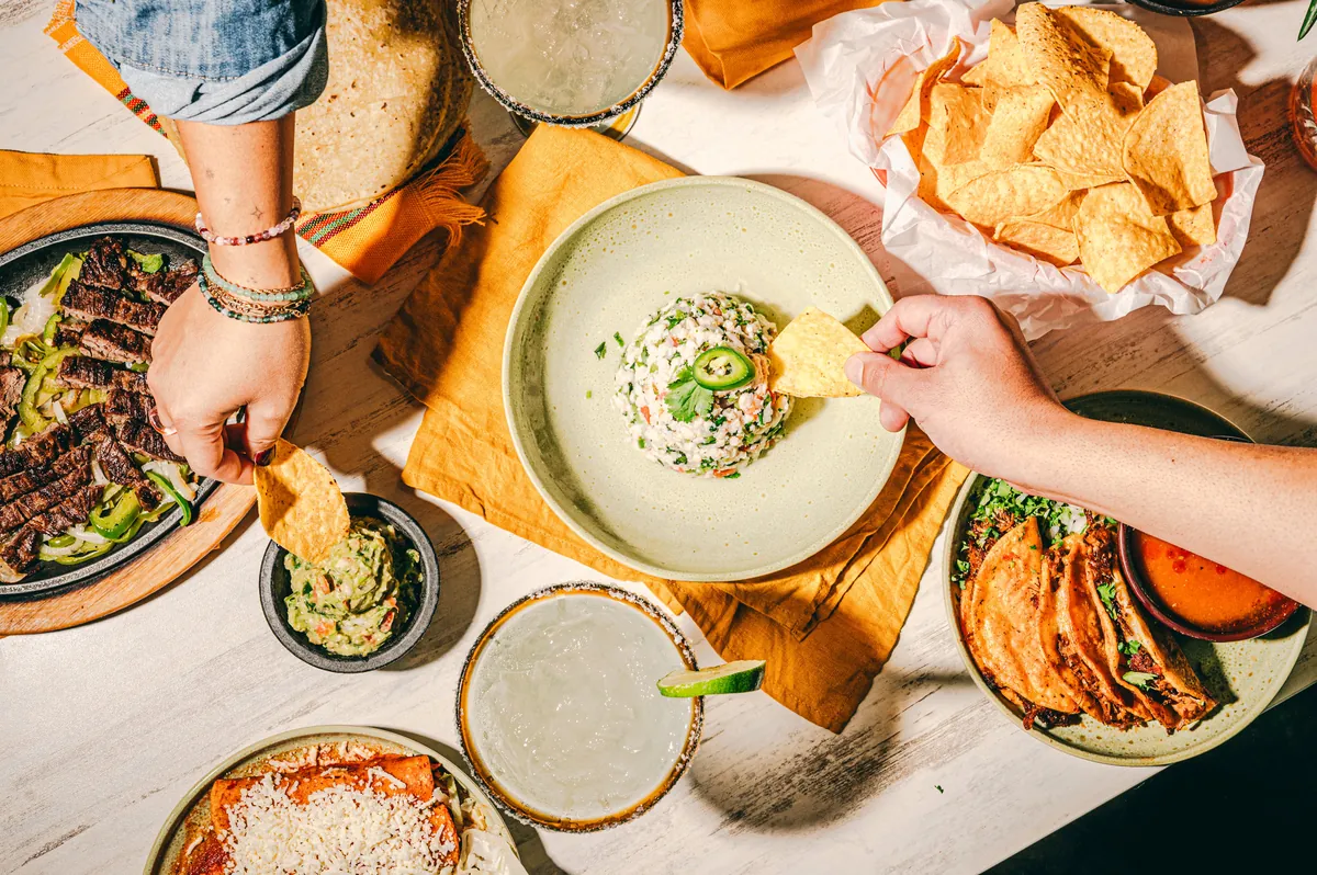 Hands reaching across a table for chips and guacamole, with enchiladas and margaritas